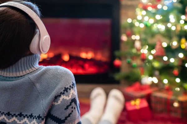 Yanlız üzgün kulaklıklı kadın kış akşamı şömine ateşi ve yılbaşı ağacının yanında oturup ısınıyor..