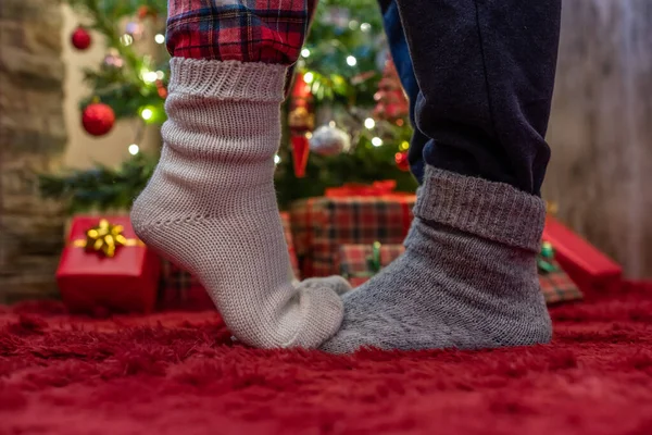 Woman feet standing in tip toe in winter socks on male lags on a fluffy red blanket near a Christmas tree with gifts. Concept