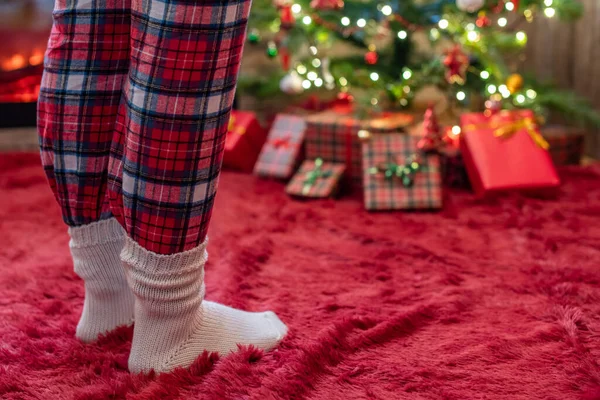 Female feet in pajama and winter socks standing near a Christmas tree with gifts and fireplace at morning. Concept