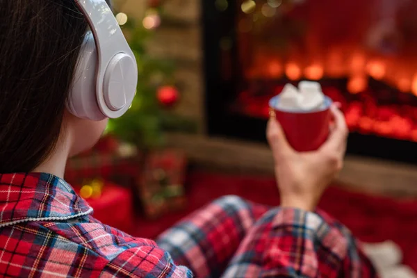 冬天的晚上 穿着睡衣 头戴耳机 拿着热可可和棉花糖的女人坐在壁炉边和圣诞树旁取暖 — 图库照片