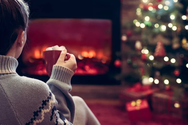 暖炉の炎とクリスマスツリーの近くの冬の夜に座って温暖化ホットココアとマシュマロのカップを持つ女性 — ストック写真