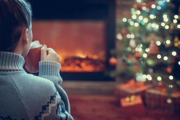 Elinde bir fincan sıcak kakao ve lokum olan kadın kış akşamı şömine ateşi ve noel ağacının yanında oturup ısınıyor..