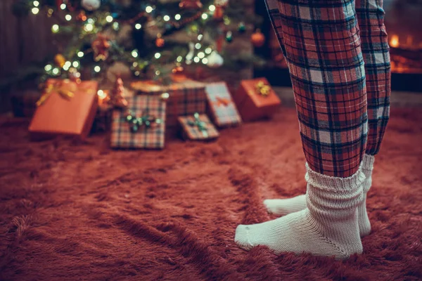 Pijama giymiş kadın ayakları ve kış çorapları Noel ağacının yanında duruyor. Hediyeler ve şömineyle birlikte. Kavram