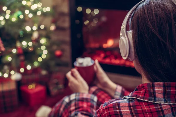 冬天的晚上 穿着睡衣 头戴耳机 拿着热可可和棉花糖的女人坐在壁炉边和圣诞树旁取暖 — 图库照片