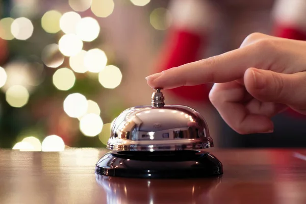 Hand of guest ringing reception bell on desk of guesthouse, hotel at christmas time. Color shining garland on christmas tree on background. Travel concept.