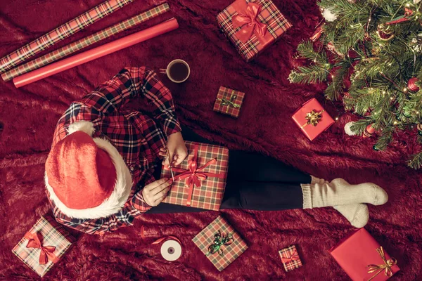 Dizlerinin üzerinde Noel hediyesi kutusu olan bir kadının en iyi görüntüsü. Noel ağacının yanındaki kabarık ekose üzerinde oturan aile için kadın paketleme hediyeleri. Düz yatıyordu.