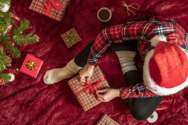 クリスマスツリーの近くで遊ぶふわふわの上に座っている家族のためのクリスマスプレゼント箱を包んだ赤いサンタクラスの帽子の女性のトップビュー — ストック写真