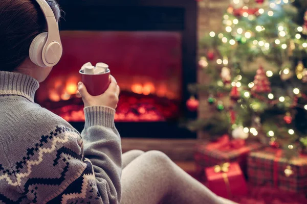 冬天的夜晚 站在壁炉边的火焰和圣诞树旁 戴着耳机 拿着一杯热可可和棉花糖的女人坐着取暖 — 图库照片