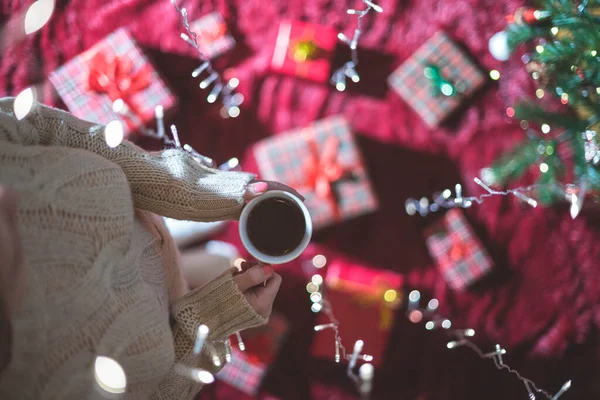 Elinde bir fincan sıcak çikolata ve aile için Noel hediyelerini Noel ağacının yanında yumuşak ekose kumaşla paketleyen bir kadın. Parlayan çelenk boyunca en üst manzara.