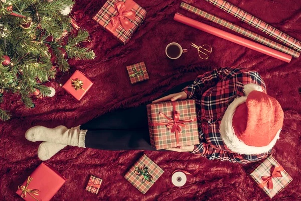 Dizlerinin üzerinde Noel hediyesi kutusu olan bir kadının en iyi görüntüsü. Noel ağacının yanındaki kabarık ekose üzerinde oturan aile için kadın paketleme hediyeleri. Düz yatıyordu.