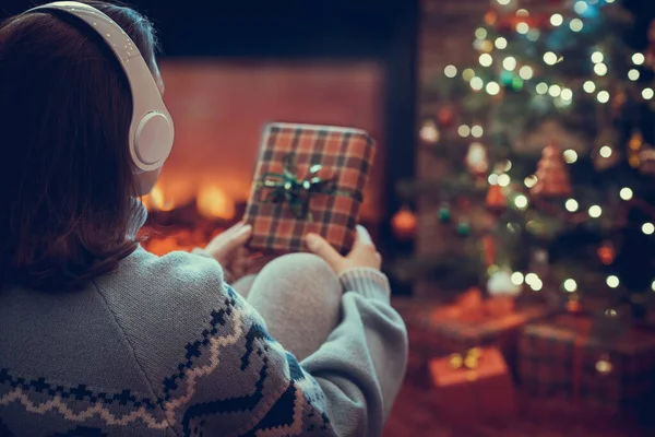 女性でヘッドフォンでクリスマスギフトボックスで手に座ってふわふわ暖炉とクリスマスツリーの近くで再生 — ストック写真