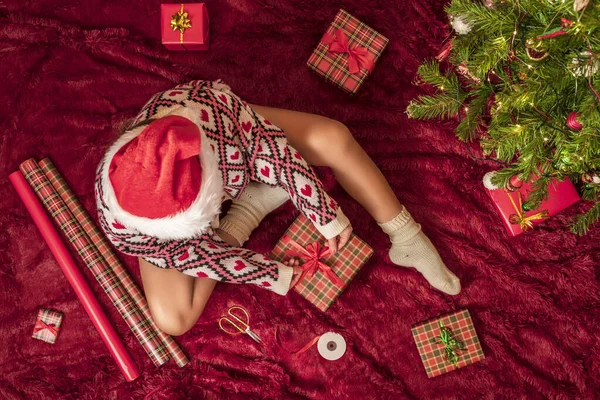 Kırmızı Noel Baba şapkalı bir kadının, Noel ağacının yanındaki kabarık ekose kumaşta oturan aile için Noel hediyesi kutularını paketlediği en iyi manzara. Düz yatıyordu.