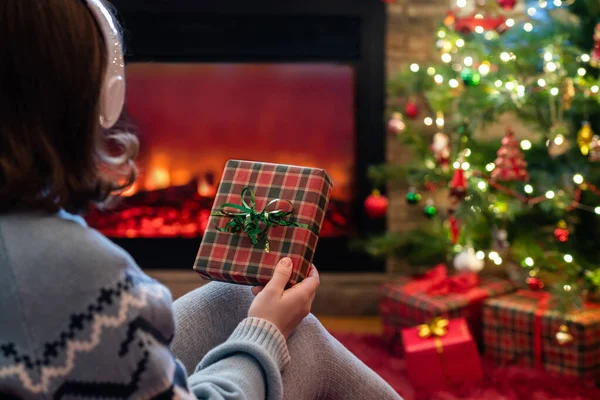头戴耳机 手里拿着圣诞礼物盒的女人坐在壁炉边蓬松的格子花上和圣诞树旁 — 图库照片