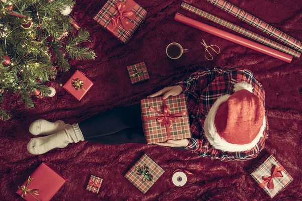膝の上にクリスマスギフトボックスを持つ女性のトップビュー クリスマスツリーの近くで遊ぶふわふわの上に座っている家族のための女性の包装ギフト — ストック写真