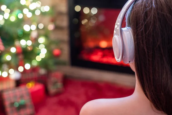 暖炉の炎とクリスマスツリーの近くの冬の夜に座って暖まるヘッドフォンで一人で悲しい女性 — ストック写真