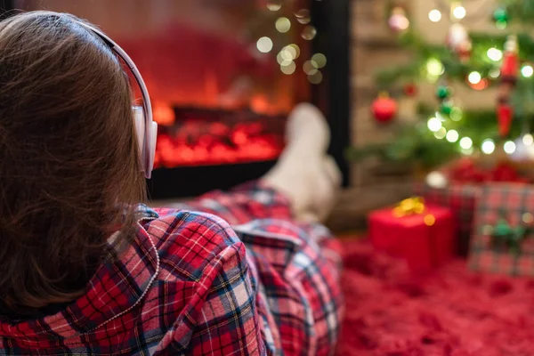 冬天的傍晚 穿着睡衣 头戴耳机的女人躺在壁炉边 听着音乐 暖烘烘地听着炉边的火焰圣诞树 — 图库照片