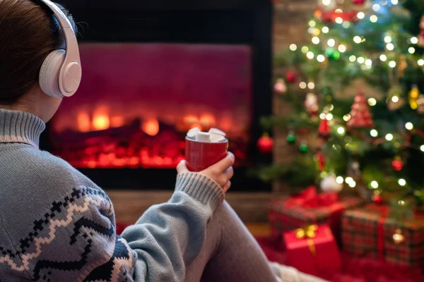 冬天的夜晚 站在壁炉边的火焰和圣诞树旁 戴着耳机 拿着一杯热可可和棉花糖的女人坐着取暖 — 图库照片