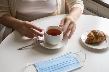 Kadın ziyaretçi bir fincan kahve ve kruvasanla kahvaltı yapıyor. Kafenin penceresinin yanındaki masaya tıbbi koruyucu yüz maskesi koyuyor. Yukarıdan görüntüle.