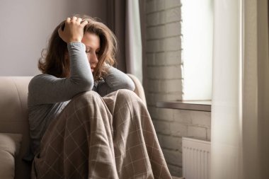 Üzgün, korkmuş, panik atak geçiren bir kadın kafasını eliyle tutuyor. Ekoseye sarılı yalnız bir kız. Dairelerin penceresinin yanında oturuyor..