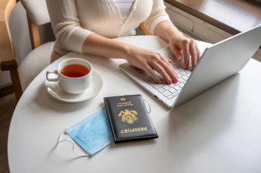 Bir fincan kahve ve ayçöreği olan kadın turist, tıbbi koruyucu yüz maskesi ve Amerikan pasaportunu havaalanındaki kafenin penceresinin yanındaki masaya koyuyor. Coronavirus sırasında seyahat kavramı