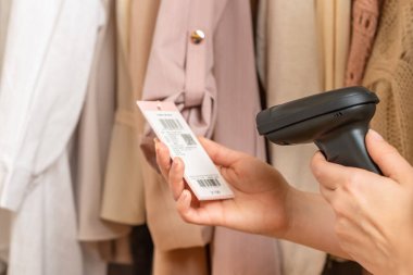 Bir depo çalışanı barkod tarayıcısı kullanarak Çin kıyafetlerini kabul ediyor. Kadın ceketinin fiyat etiketinden barkod okuyor.  