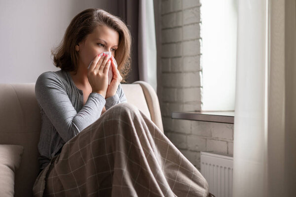 Sick brunette woman sneezing in a tissue sitting on the couch in the living room near window