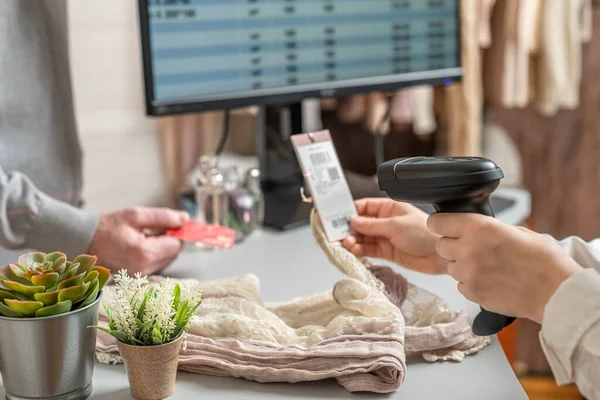 Man with credit card buying gift in a female clothing store. Woman cashier scanning price tag using barcode scanner at the checkout.