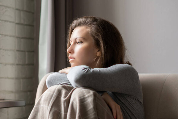 Portrait of lonely sad caucasian young woman wrapped in a plaid sitting near window of apartments.