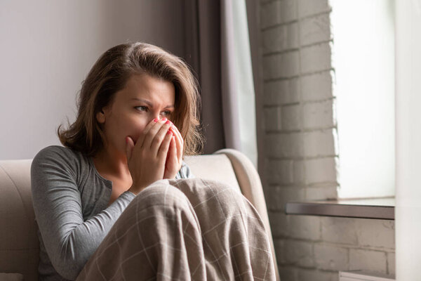 Sick brunette woman sneezing sitting on the couch in the living room near window
