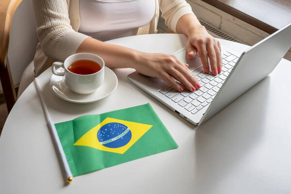 Lonely woman freelancer with flag of  Brazil enjoying having breakfast with cup of coffee working on laptop sitting near window in cafe at morning. 