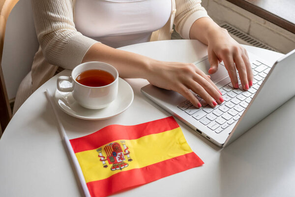 Lonely woman freelancer with flag of Spain enjoying having breakfast with cup of coffee working on laptop sitting near window in cafe at morning. 