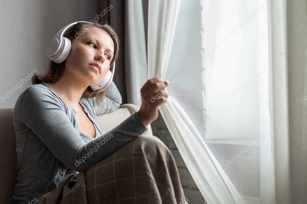 Portrait of caucasian young woman in headphones listening music sitting on sofa covered blanket and looking at window opening curtain