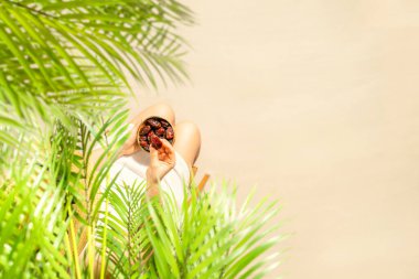 Hasır şapkalı bir kadın hindistan cevizi palmiyesi dallarının altında oturmuş elinde kraliyet meyveleri kumsaldaki bir kasede tutuyordu. Yaz. Üst görünüm