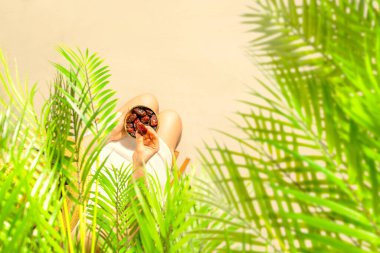 Hasır şapkalı bir kadın hindistan cevizi palmiyesi dallarının altında oturmuş elinde kraliyet meyveleri kumsaldaki bir kasede tutuyordu. Yaz. Üst görünüm