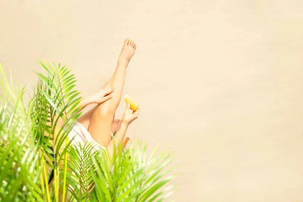 Alone woman applying sunscreen on skin sitting under palm tree branches. Female relaxation on the sand of the beach at summer vacation. Top view