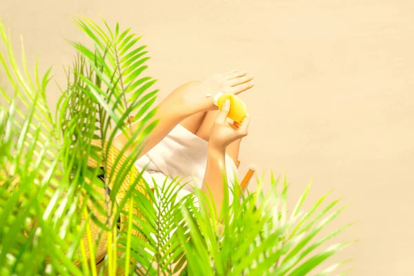 Alone woman applying sunscreen on skin sitting under palm tree branches. Female relaxation on the sand of the beach at summer vacation. Top view