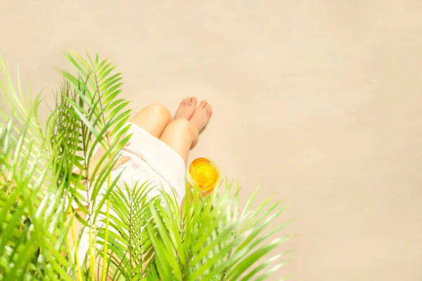 Alone woman sitting under palm tree branches with glass of water with piece orange. Female relaxation on the sand of the beach at summer vacation. Top view