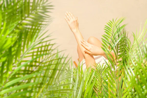 Alone woman applying sunscreen on skin sitting under palm tree branches. Female relaxation on the sand of the beach at summer vacation. Top view