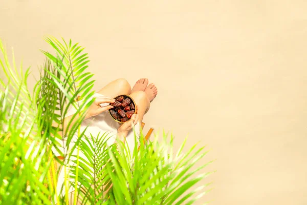 手に持っているヤシの木の枝の下に座っているわら帽子の女性は ビーチの砂の上にボウルに果物をデートします トップ表示 — ストック写真