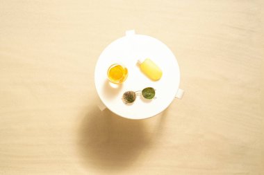 Koruma için güneş kremi, portakal suyu, kumlu sahildeki beyaz masada güneş gözlüğü. Yaz tatili. Üst görünüm