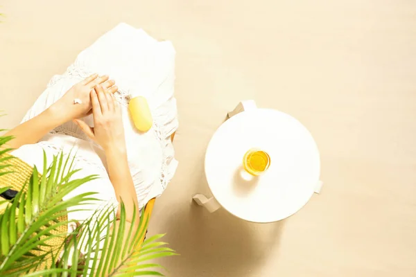 Alone woman applying sunscreen for protection skin sitting under palm tree branches near beach table with orange juice. Top view. Summer