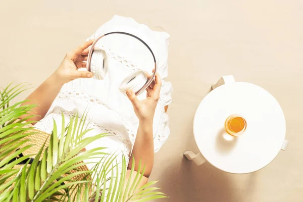 Alone woman sitting under palm tree branches. Glass of orange juice and headphones on the table. Female relaxation on the sandy beach at summer vacation. Top view