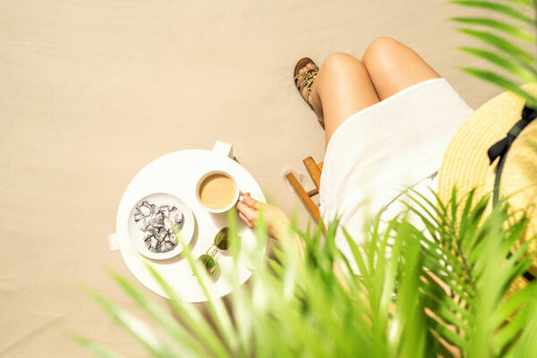 Woman on the beach drinking hot coffee with chocolate cookies sitting under palm trees at table and enjoying during exotic summer vacation. Outdoors cafe. Top view