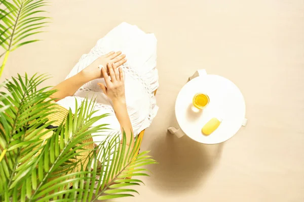 Alone woman applying sunscreen for protection skin sitting under palm tree branches near beach table with orange juice. Top view. Summer