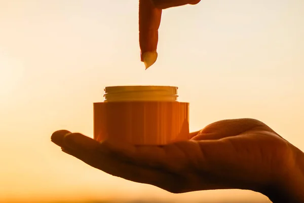 woman applying night hand cream in a jar standing near window at the evening on the sunset background. Concept.