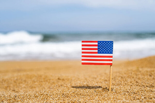 Flag of America in the form of a toothpick in the sand of beach opposite sea wave. Travel concept