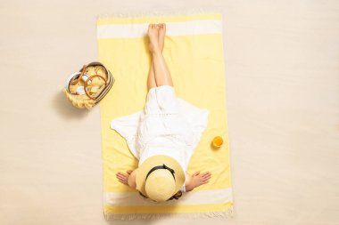 Sarı plaj havlusunda çantalı, kulaklıklı ve portakal suyu içmiş, saman şapkalı ve elbiseli yalnız bir kadın. Yaz tatili. Üst, hava görünümü