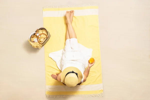 Alone woman in straw hat and dress sitting on yellow beach towel with bag, headphones, and orange juice. Summer vacation. Top, aerial view