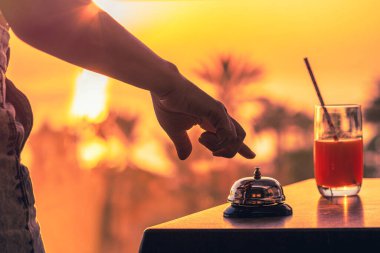 Gün batımında içki, deniz ve palmiye ağacı manzaralı otel servisi zilini çalan kadın. Seyahat konsepti. 24 saat otel resepsiyonu. Geç çıkış..
