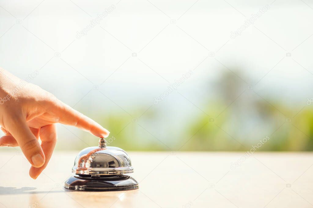 Woman ringing hotel bell of summer beach hotel front desk. Coastline sea and palm tree view. Travel concept. 
